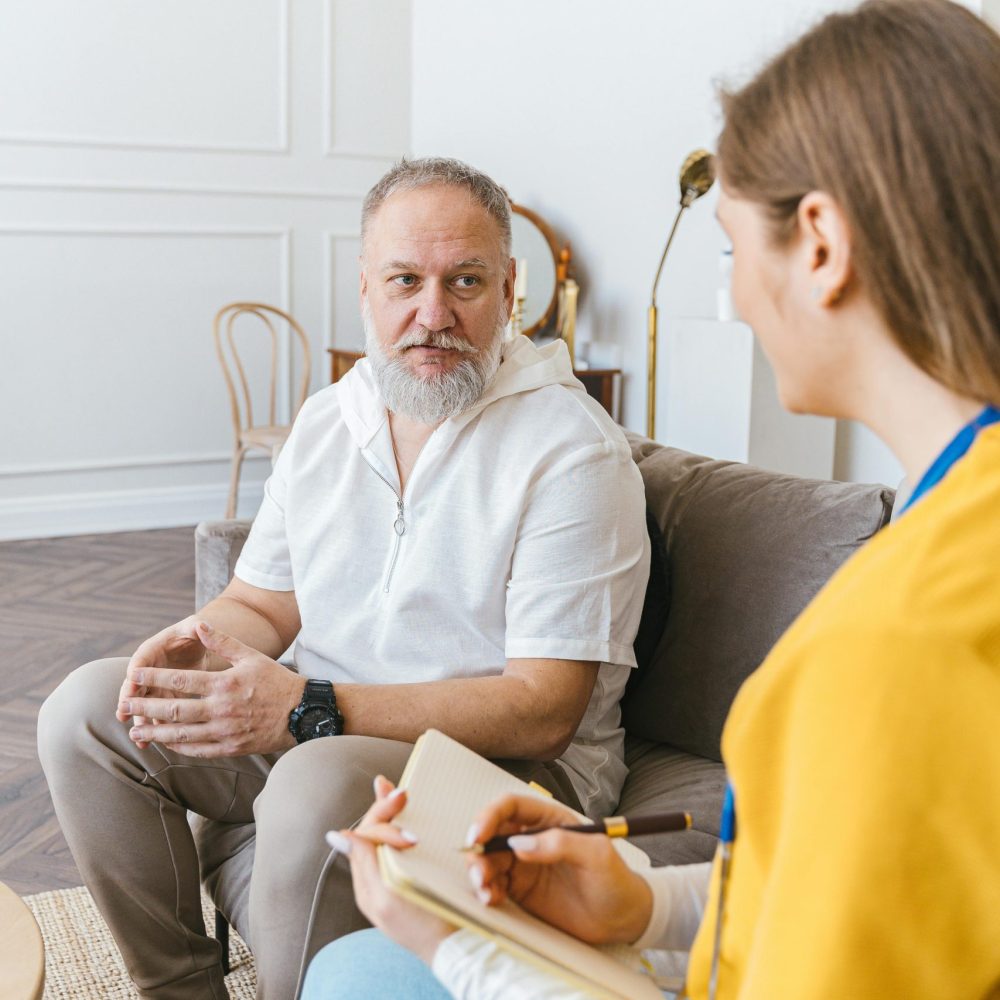 A senior man in a white shirt talks to a woman volunteer taking notes indoors.